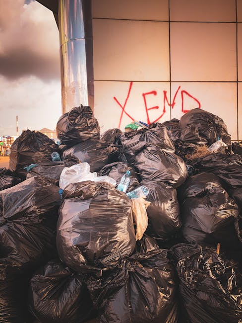 A collection of tightly packed plastic rubbish bags, primarily in yellow, white, and transparent colors, stacked against a white plastered exterior wall on a cobblestone street. The bags contain vario